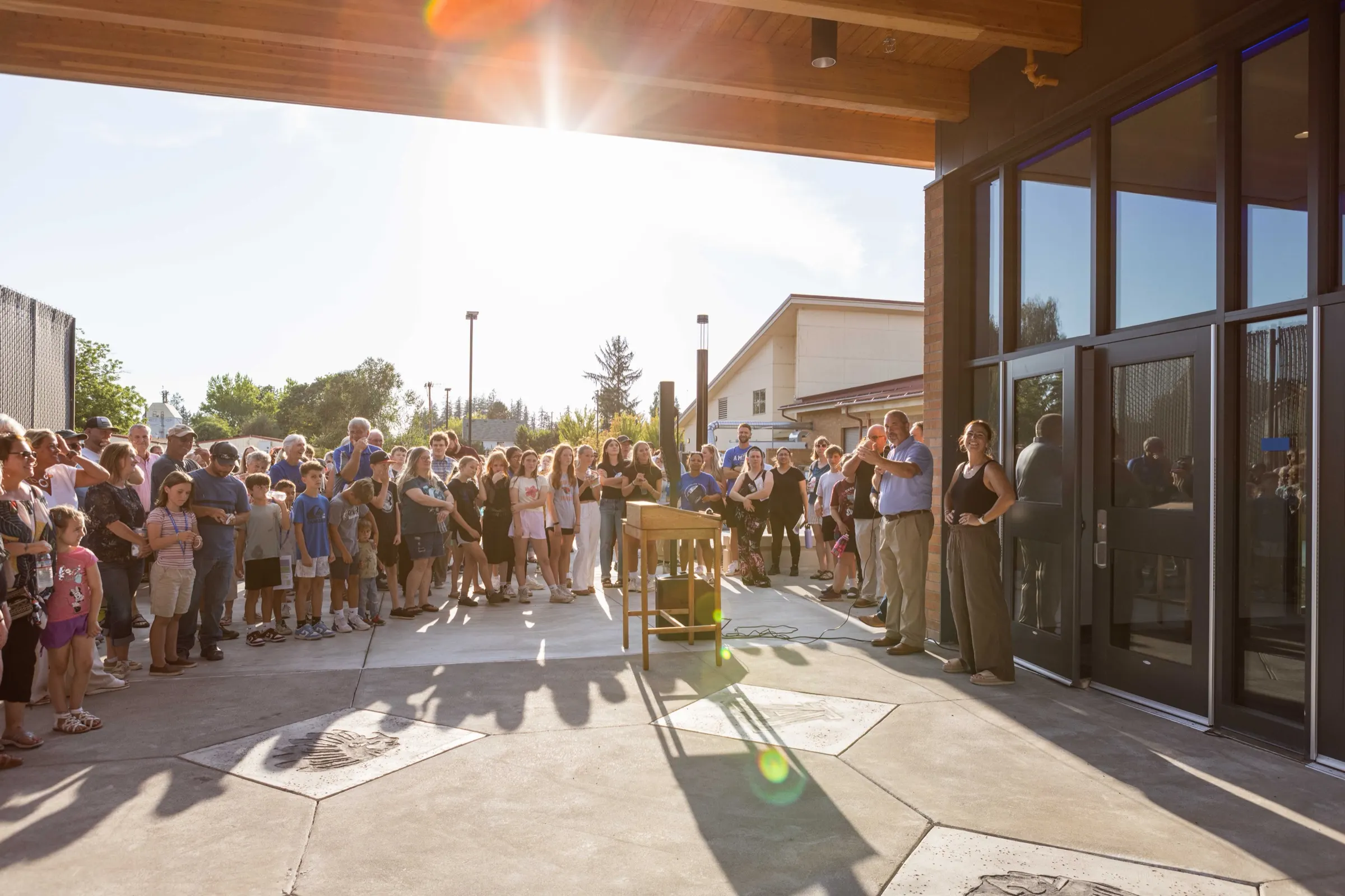 A crowd of families and children gathers outside the glass doors of a new school building for its opening ceremony on a sunny evening.