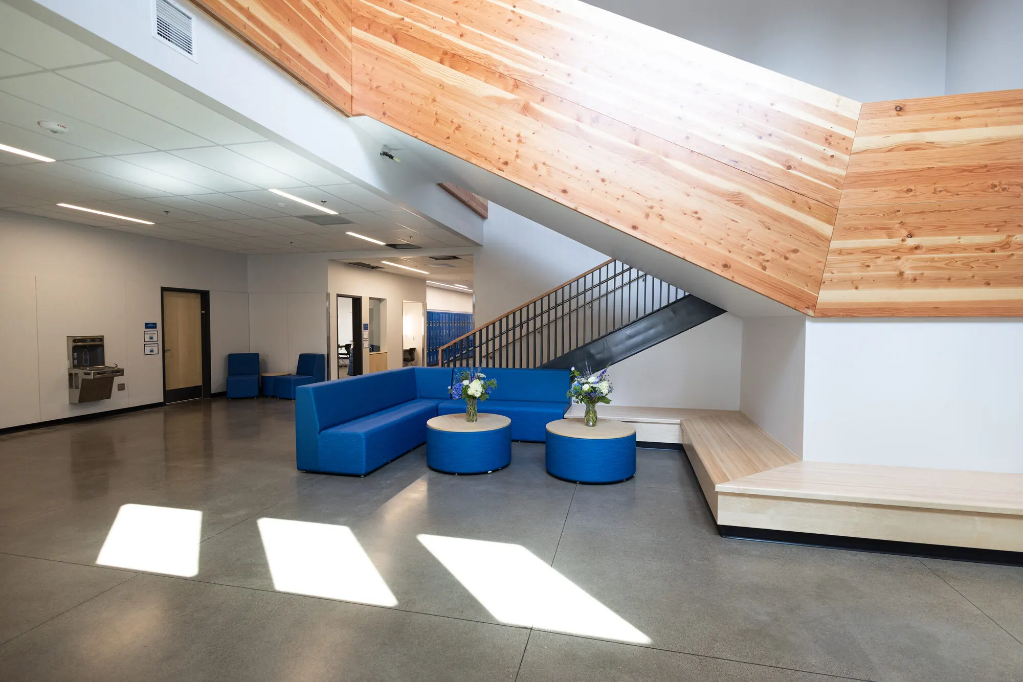 A sunlit commons at Amity Middle School with blue sofas and round tables beneath a cedar-clad staircase.