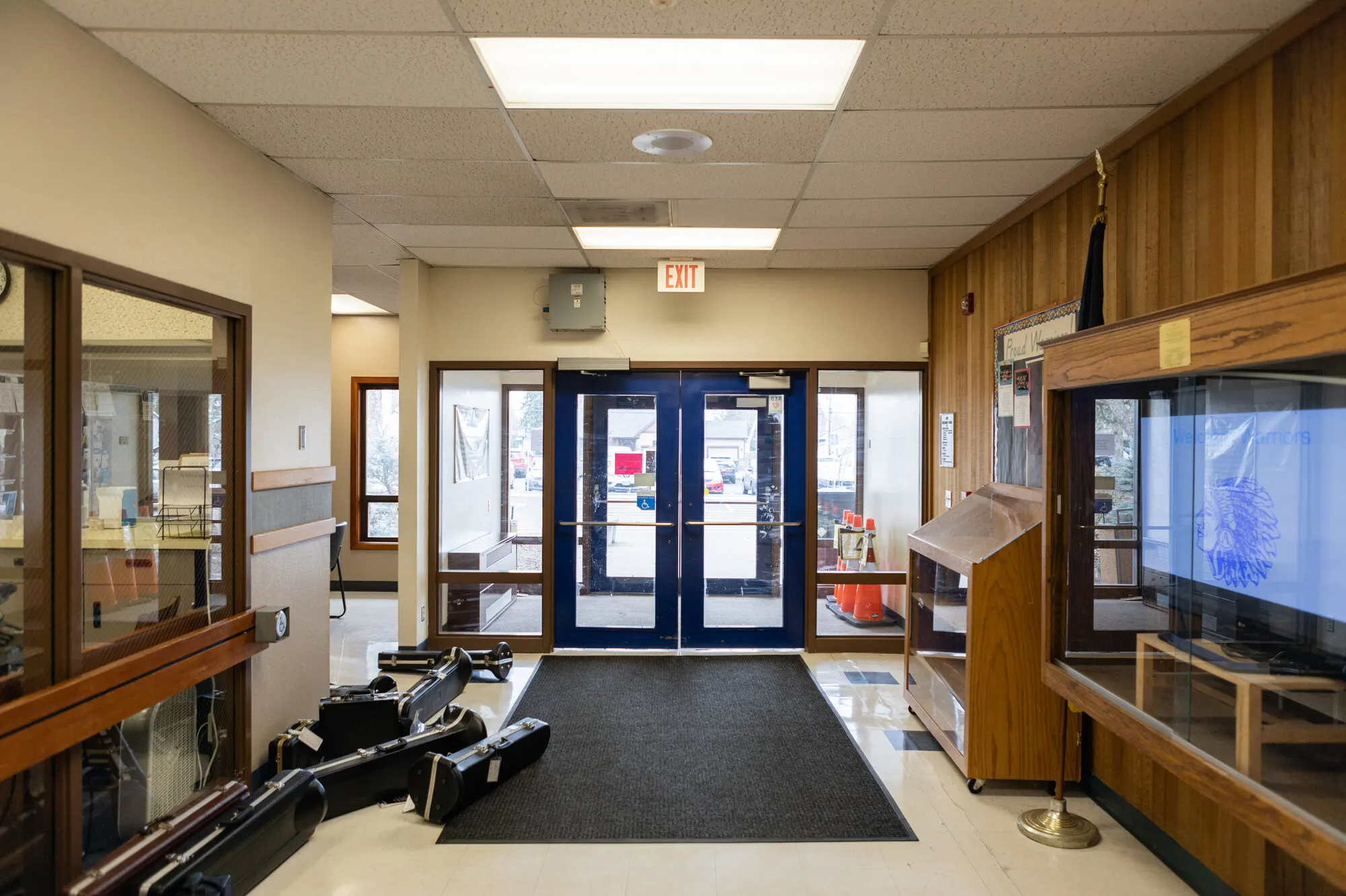 The entry hall at Amity Elementary School: blue double doors, instrument cases on the floor, and a wood-paneled wall with a glass display case.